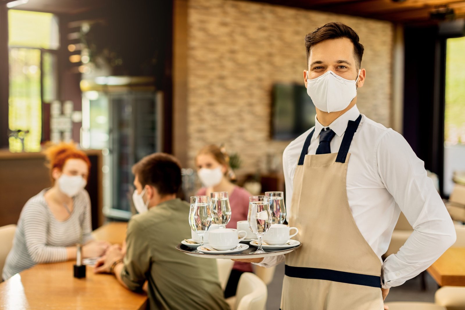Portrait of happy waiter with protective face mask serving customers in a cafe and looking at camera.