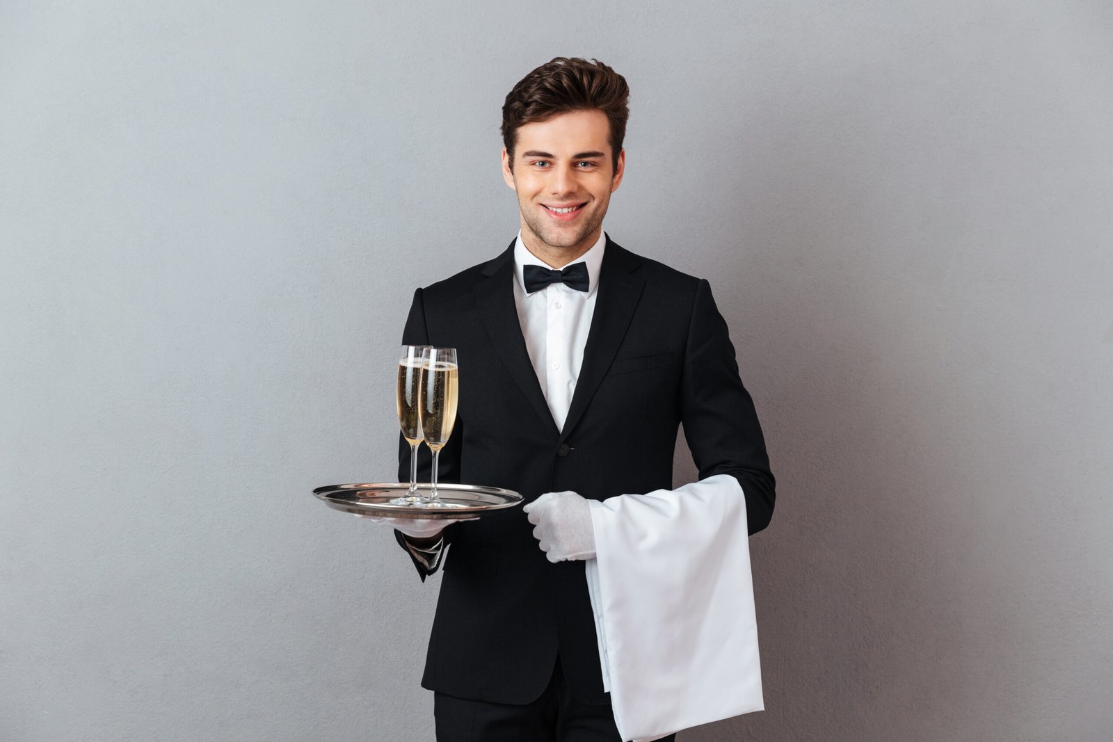 Photo of happy young waiter standing isolated over grey wall background. Looking camera holding glass of champagne and towel.